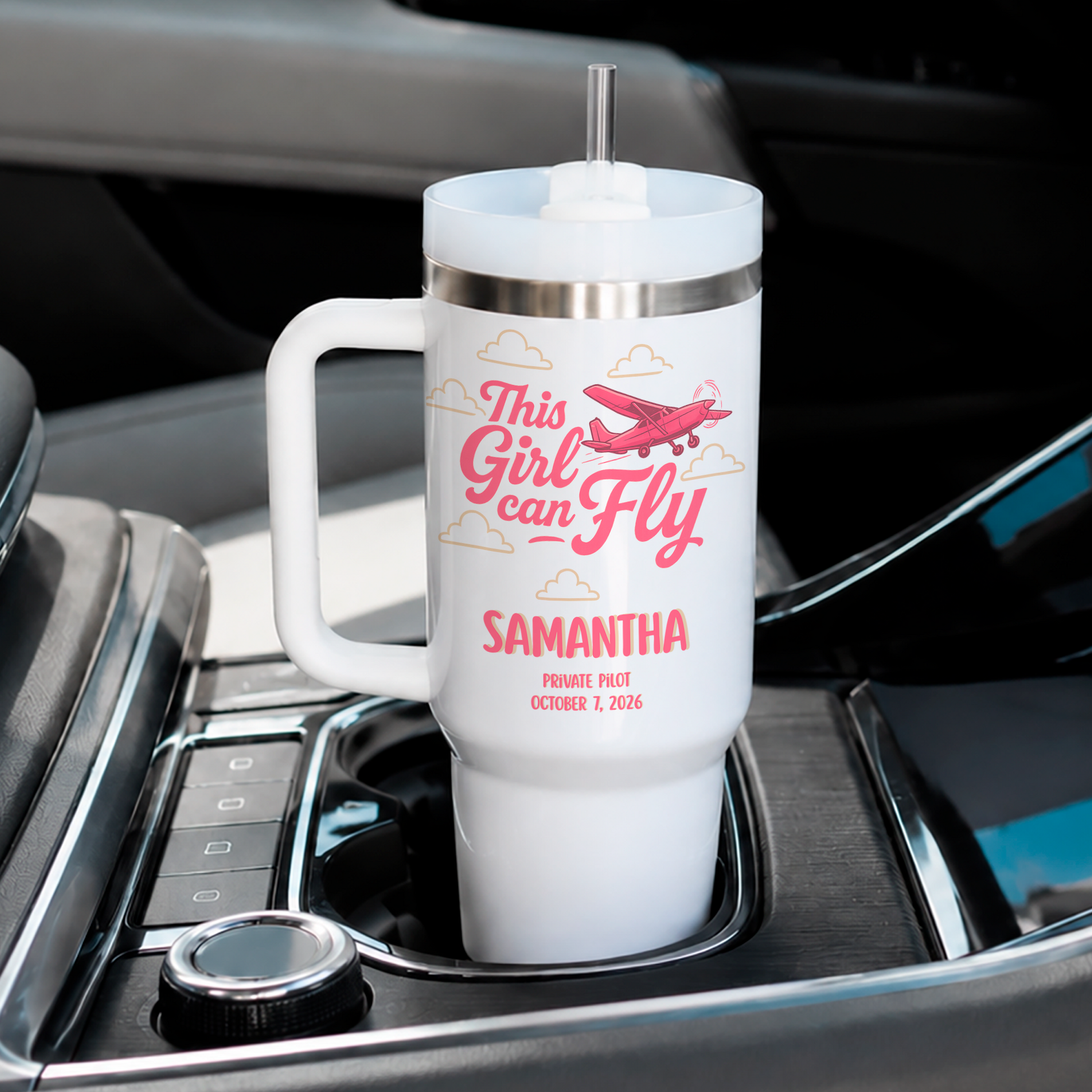 A white handled travel tumbler with a clear lid and straw sits in a car cup holder. The tumbler has a pink graphic reading 'This Girl Can Fly' with an airplane and the name 'SAMANTHA' in pink text below.