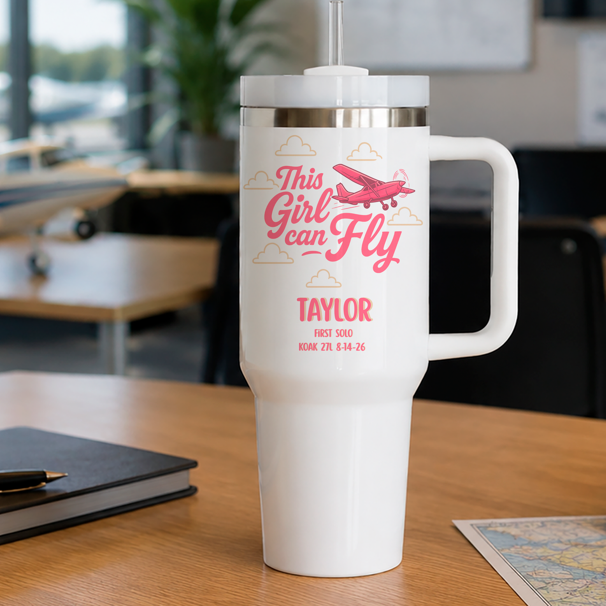 A white insulated travel tumbler with a handle and lid sits on a wooden desk in a blurred office or classroom setting. The tumbler has a pink printed design featuring the text 'This Girl Can Fly' with an airplane and clouds, plus the name 'TAYLOR' and dates below.