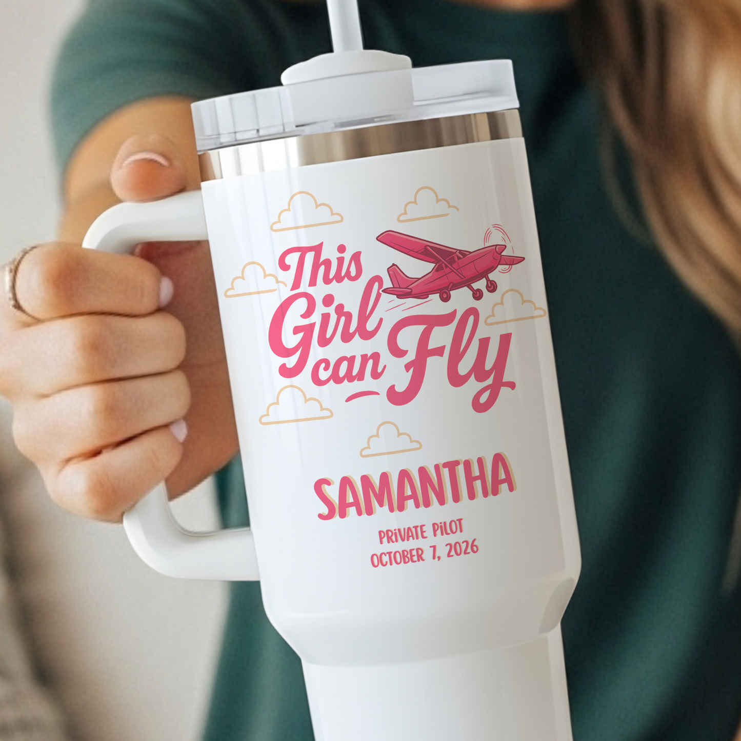 A person holds a white handled tumbler with a clear lid and straw. The tumbler is printed with pink text reading 'This Girl can Fly' and 'SAMANTHA', plus a pink airplane and small cloud illustrations.