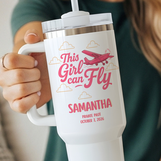 A person holds a white handled tumbler with a clear lid and straw. The tumbler is printed with pink text reading 'This Girl can Fly' and 'SAMANTHA', plus a pink airplane and small cloud illustrations.