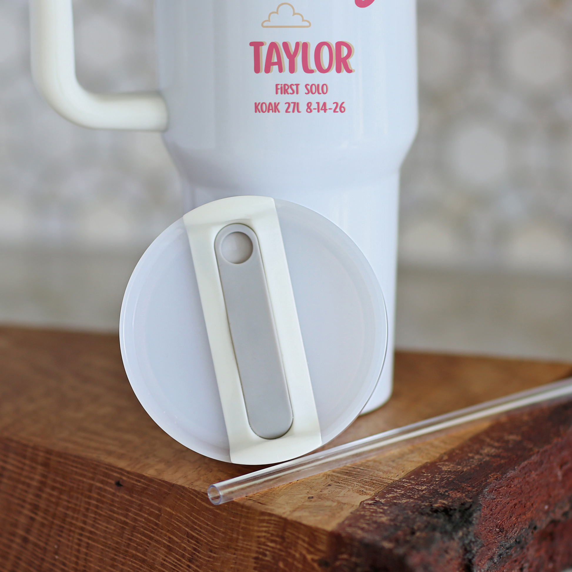 A white insulated travel mug with a handle and printed red text reading 'TAYLOR FIRST SOLO KOK 27L 8-14-26', shown with a white lid and clear straw on a rustic wooden surface.
