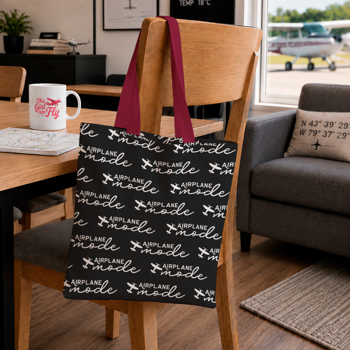 A black tote bag with white 'Airplane Mode' text and airplane icons hangs from a wooden chair in a living room setting. The bag has dark red handles and is shown in a cozy indoor environment with a table, mug, and armchair in the background.