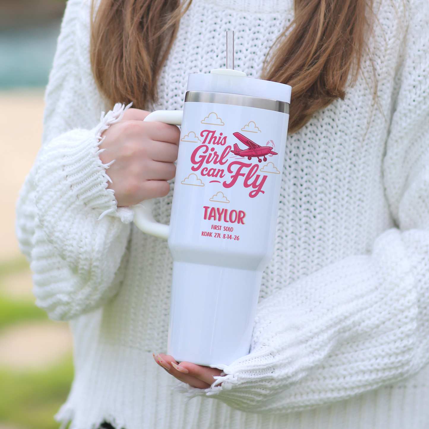 A person in a white chunky knit sweater holds a white handled tumbler with a silver lid. The tumbler is printed with pink text reading 'This Girl Can Fly' and 'TAYLOR' with a small airplane graphic.