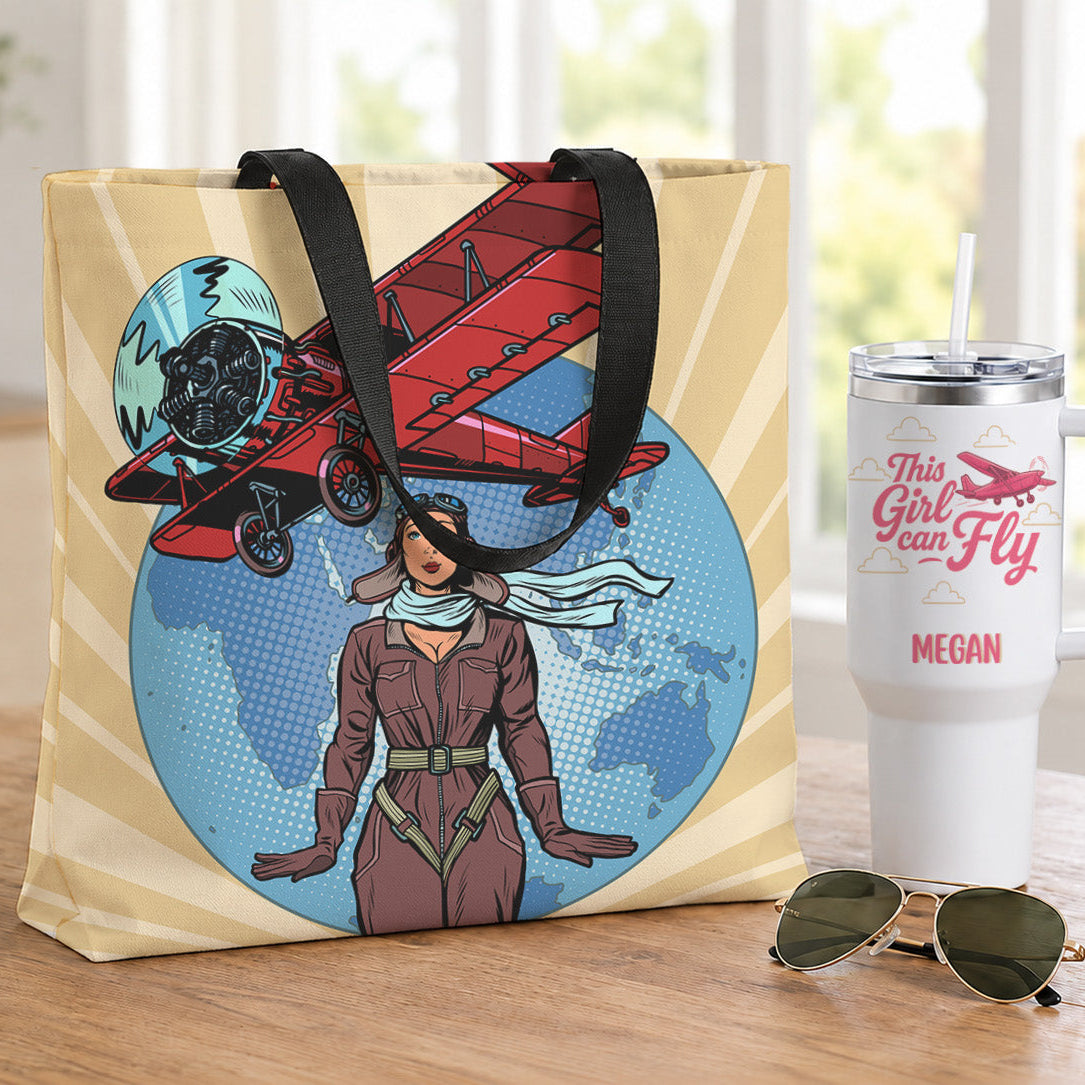 A canvas tote bag with a vintage pop-art illustration of a woman in a flight suit and a red biplane, displayed on a wooden table next to a white insulated tumbler and sunglasses.