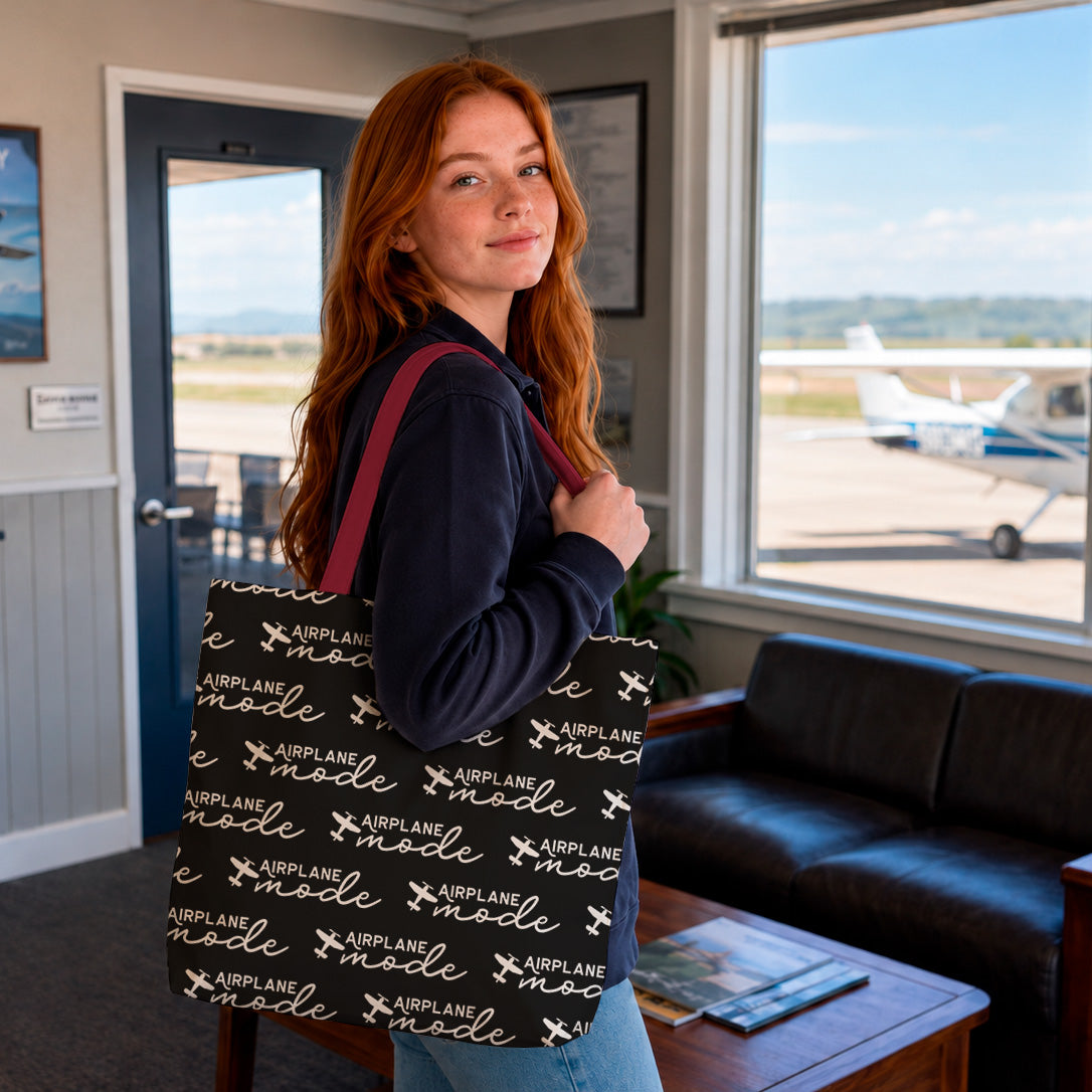 A woman with long red hair stands in an airport lounge holding a large black tote bag printed with the words 'Airline Mode' and small airplane icons. A small airplane is visible outside the window behind her.