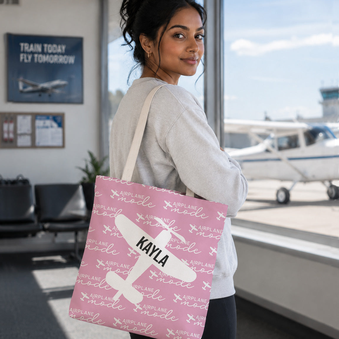 A woman standing in an airport terminal holds a pink tote bag printed with white airplane graphics and the name 'KAYLA' in black. The background shows a small airplane, windows, and airport interior.