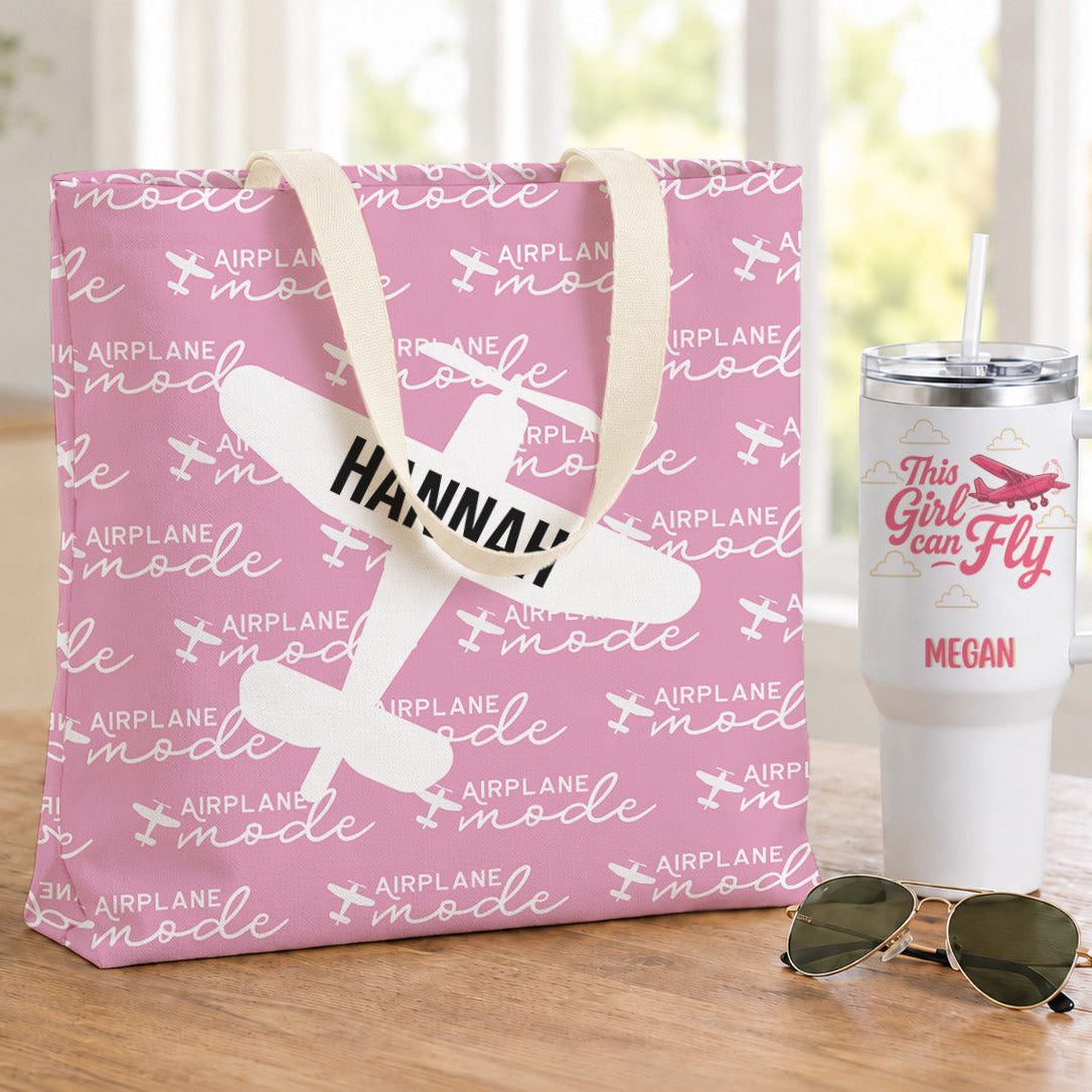 A pink gift bag and a white handled tumbler with airplane-themed custom prints are displayed on a wooden table, alongside sunglasses. The bag and tumbler feature the text 'Hannah' and 'This Girl Can Fly' with airplane graphics.