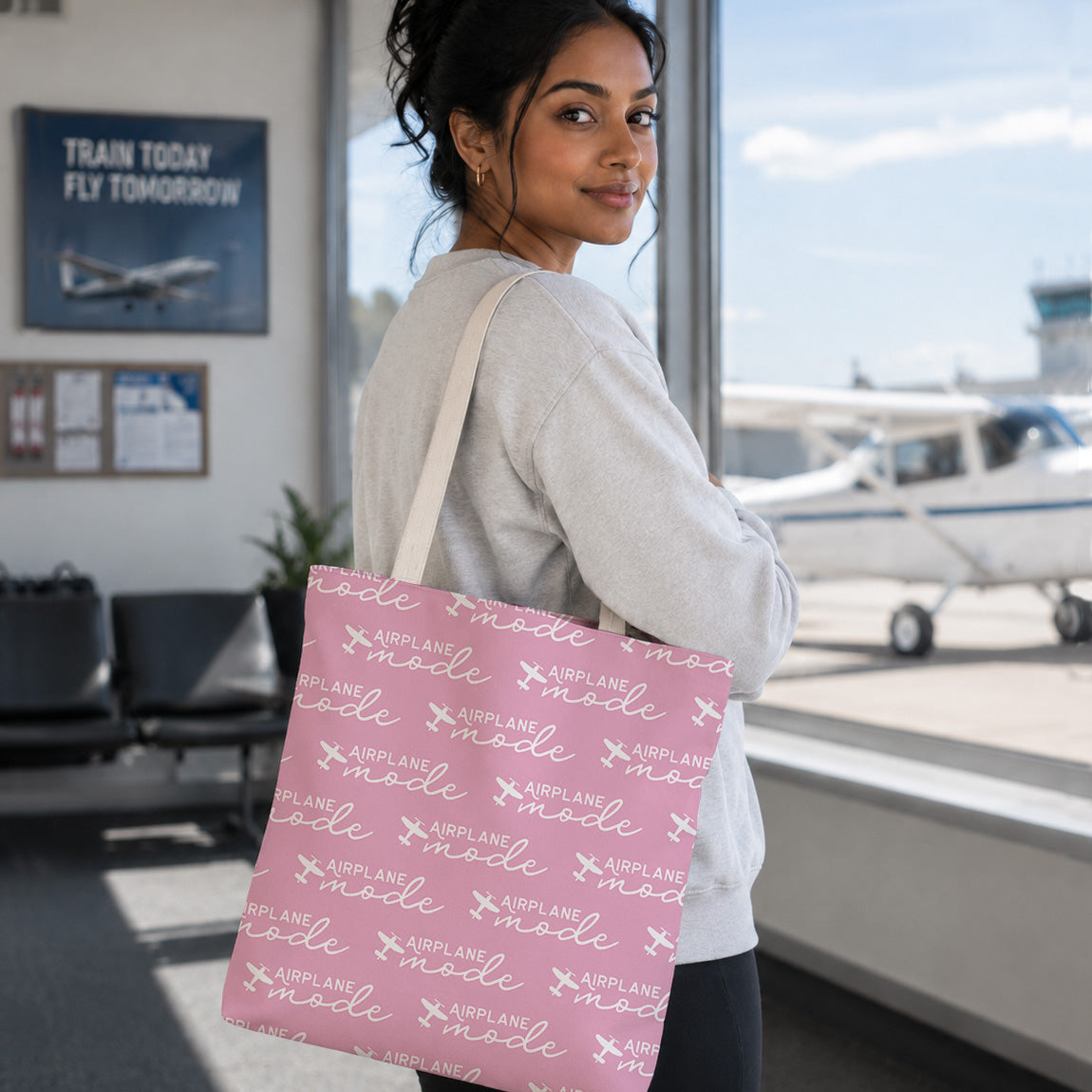A woman in a white sweatshirt carries a pink tote bag with a white airplane and 'mode' pattern, standing in an airport lounge with a small plane visible through the window.