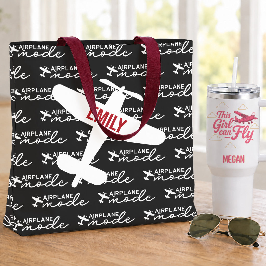 A black tote bag with a white 'Airplane Mode' pattern and red handles, featuring a white airplane graphic and the name 'EMMY' in red. It is displayed on a wooden table next to a white insulated tumbler and sunglasses.
