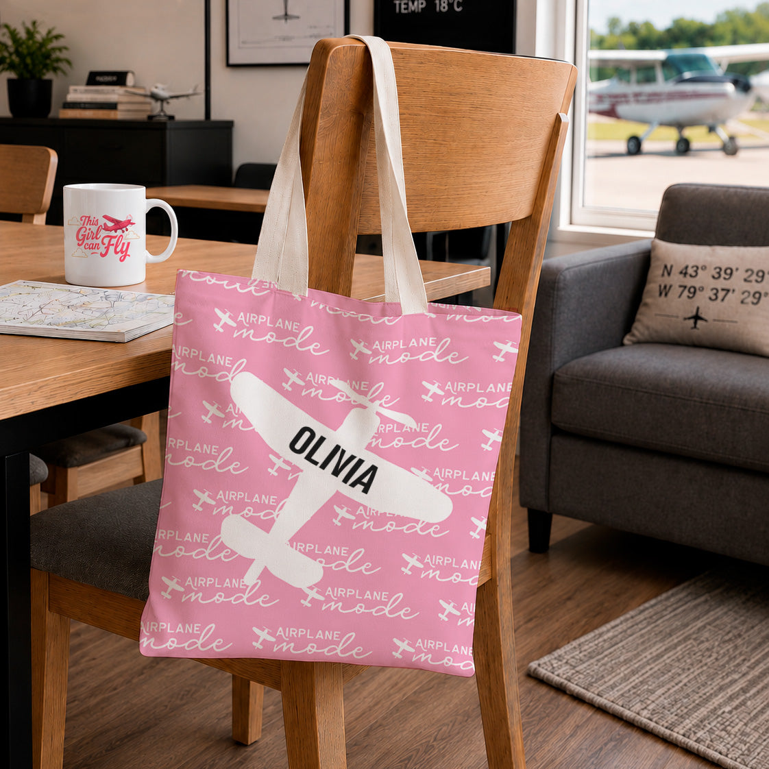 A pink fabric tote bag with a white airplane graphic and the name 'OLIVIA' printed on it, hanging on a wooden chair in a living room setting.