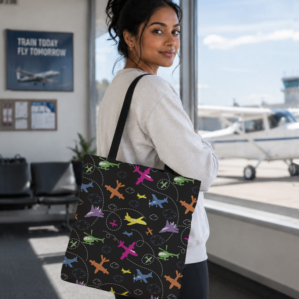 A woman in a white sweatshirt carries a black tote bag with a colorful airplane pattern, standing in an airport terminal near a window with a small plane visible outside.