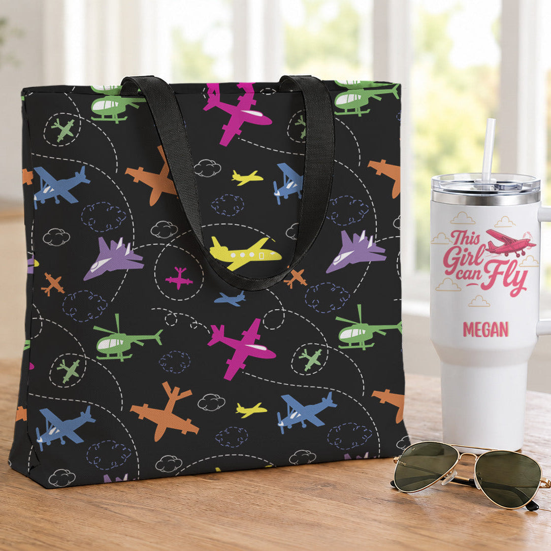 A black tote bag with a colorful airplane pattern sits on a wooden table next to a white insulated tumbler and sunglasses in a bright indoor setting.