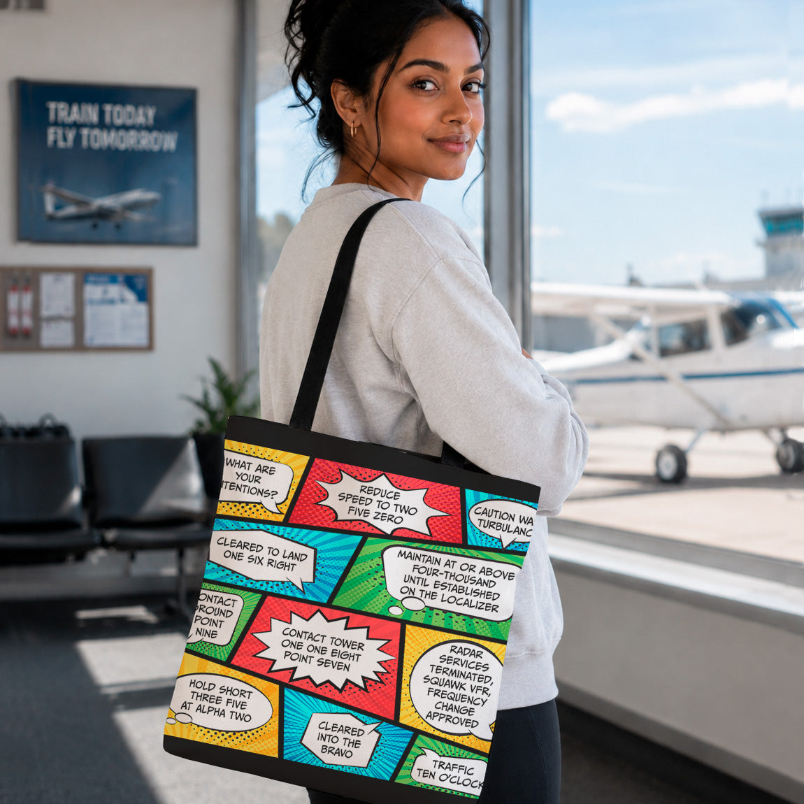 A woman in a white sweatshirt stands in an airport holding a black tote bag printed with colorful comic book speech bubbles in red, yellow, blue, green, and white.
