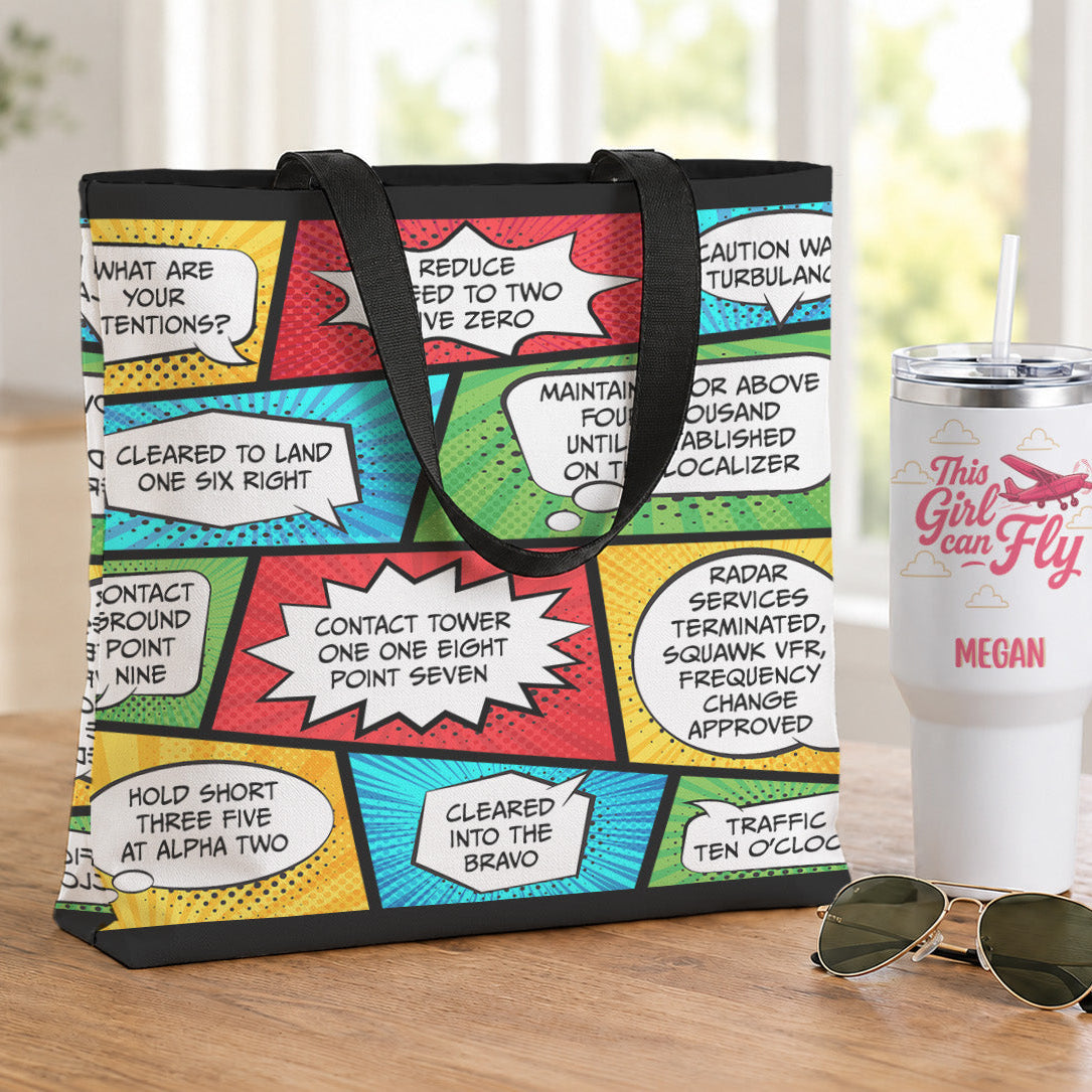 A tote bag with a colorful comic book-style print featuring text bubbles and black handles, displayed on a wooden table next to a white handled tumbler and sunglasses.