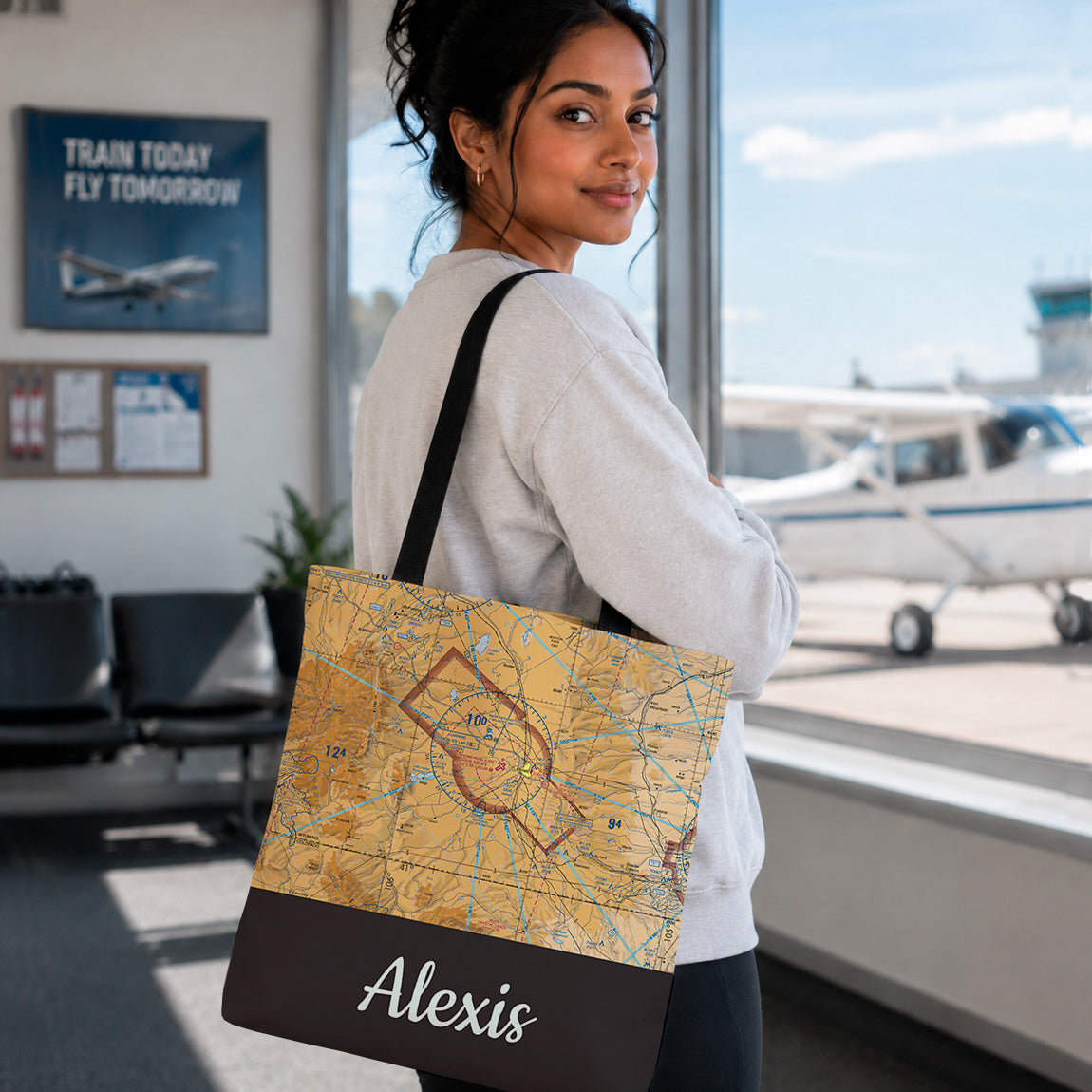 A woman standing in an airport terminal holds a tote bag with a vintage-style map print and a black bottom panel printed with the name 'Alexis' in white script.