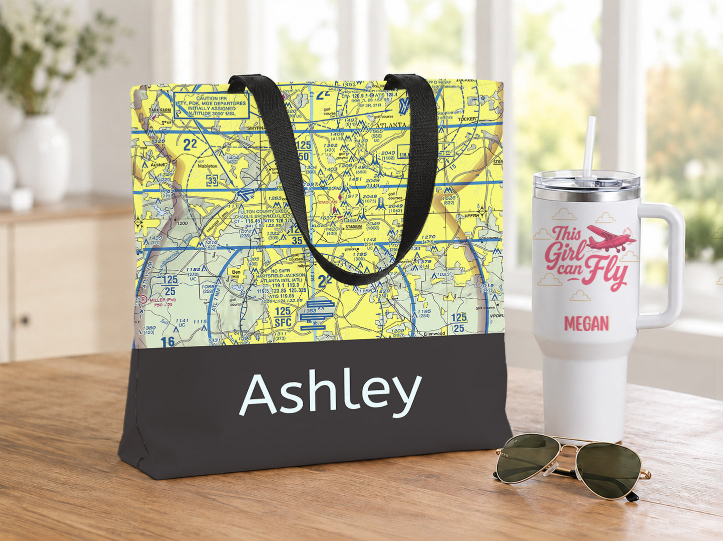 A personalized canvas tote bag with a yellow and blue nautical map print on the top half and a black bottom panel printed with the name 'Ashley' in white. The bag is styled on a wooden table next to a white insulated travel mug with pink text and sunglasses.