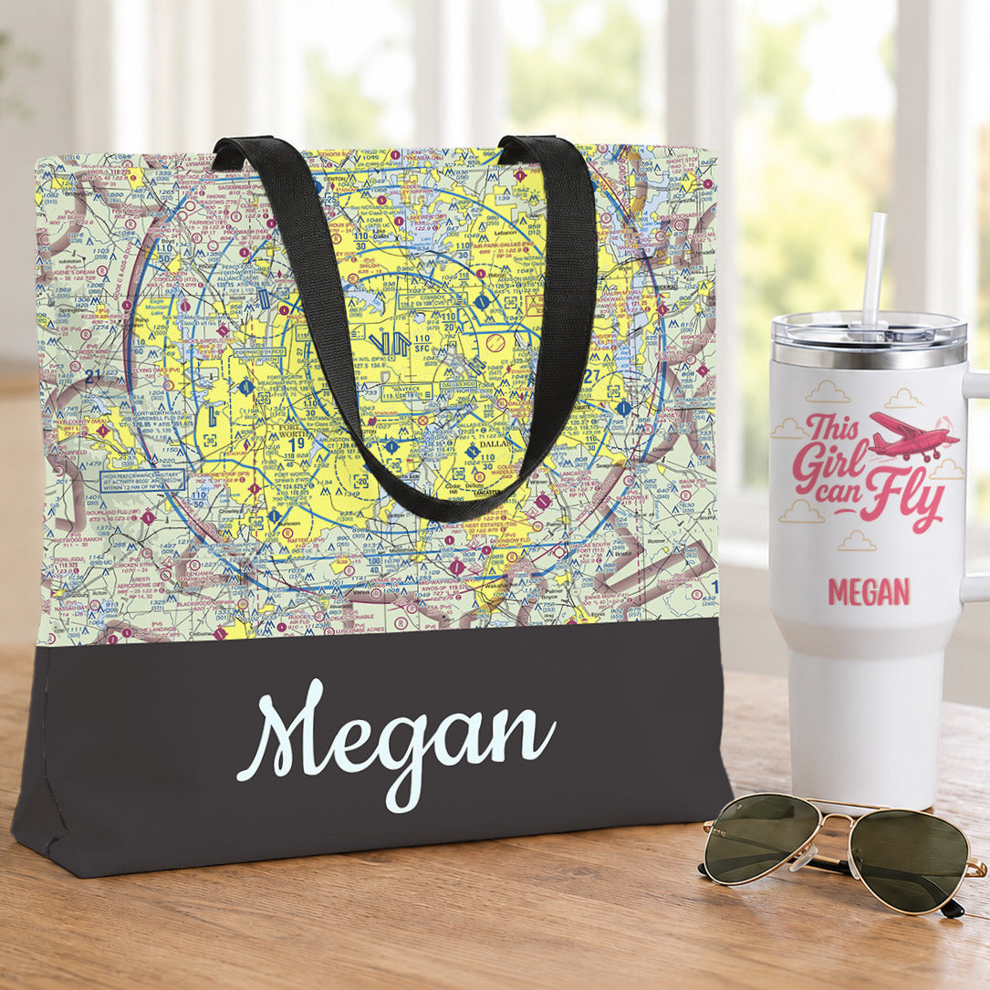 A personalized tote bag with a vintage map print and black bottom panel printed with the name Megan in white script, displayed on a wooden table next to a white insulated tumbler and sunglasses.