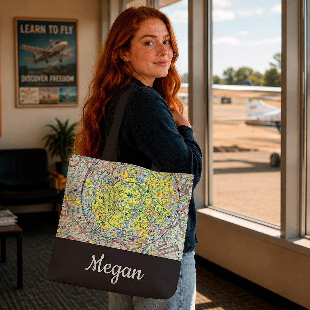A woman with red hair stands in an airport lounge holding a personalized tote bag. The bag features a colorful vintage-style map print on the upper half and a dark brown bottom panel with the name 'Megan' written in white script.