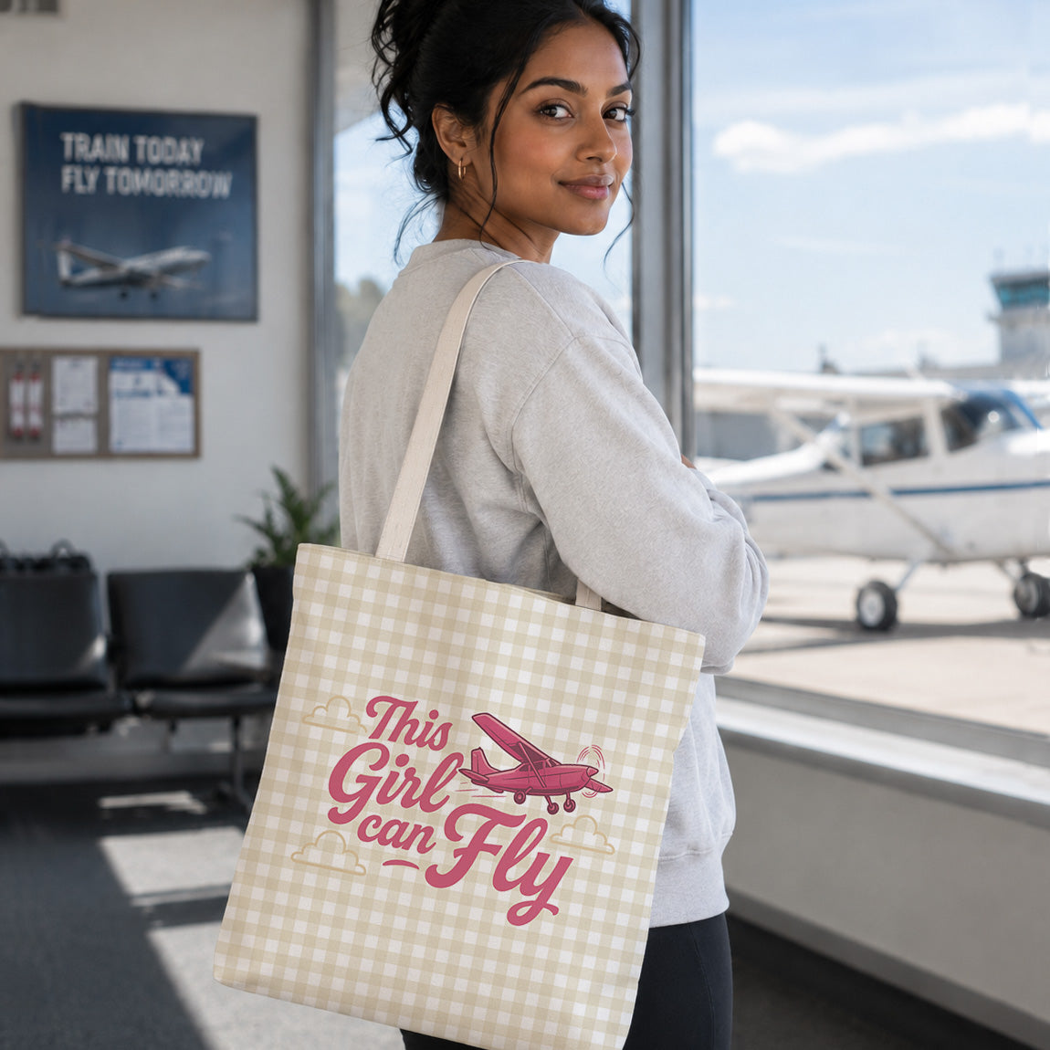 A woman in a light gray sweatshirt stands in an airport terminal holding a beige and white gingham tote bag with pink text reading 'This Girl can Fly' and a pink airplane graphic. A small white airplane is visible outside the window.