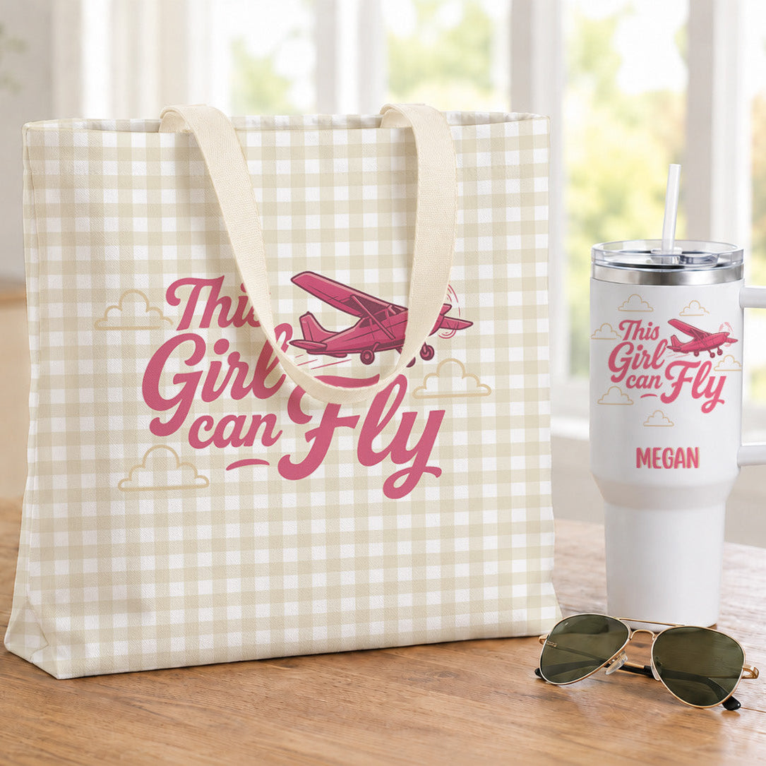 A cream and white gingham tote bag with a pink airplane graphic and the text 'This Girl Can Fly' sits on a wooden table next to a matching white insulated tumbler and sunglasses in a bright indoor setting.