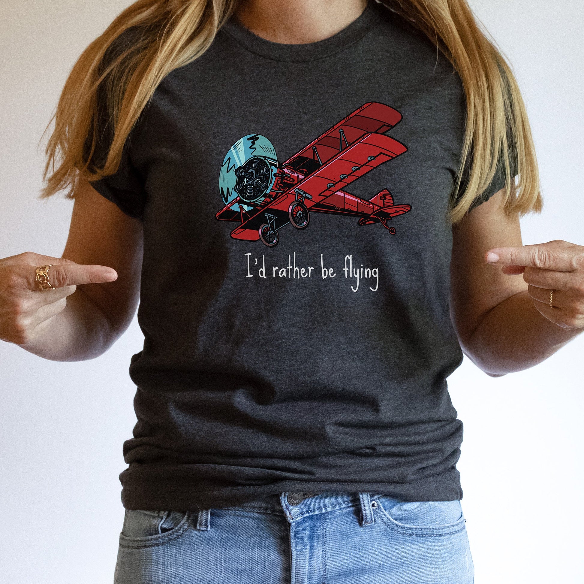 Woman pointing at the t-shirt she is wearing featuring a large red biplane and text stating I'd rather be flying underneath
