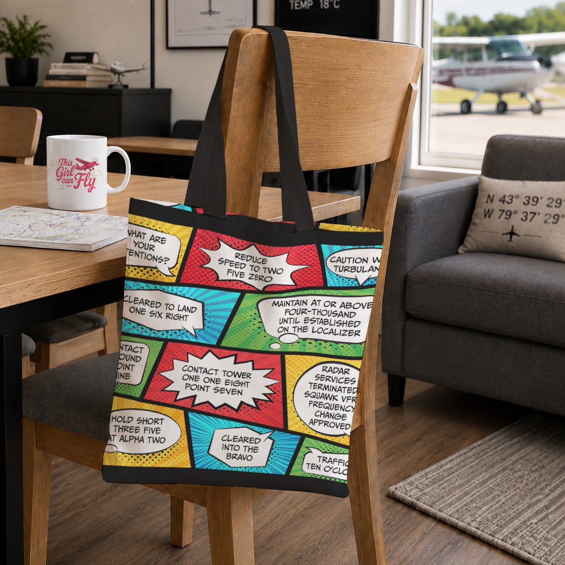 A canvas tote bag with a colorful comic book panel print and black handles, hanging on a wooden chair in a living room setting.
