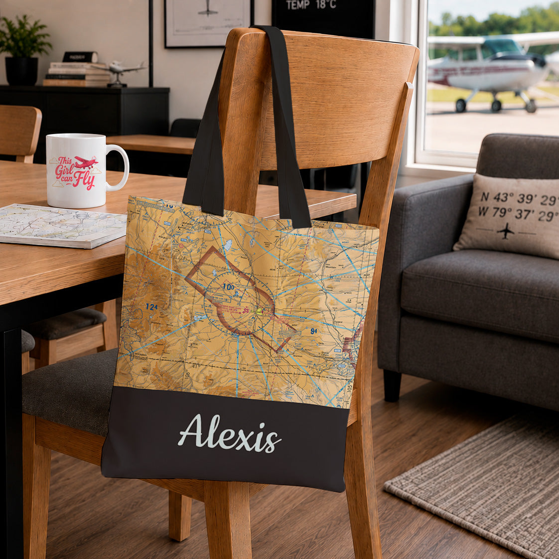 A canvas tote bag with a vintage-style map print and a dark bottom panel featuring the name 'Alexis' in white script, hanging on a wooden chair in a living room setting.